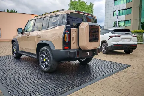 Beżowy Jetour T2 z kołem zapasowym z tyłu, obok białego SUV-a. Sprawdź Jetour T2 cena i dostępne opcje.