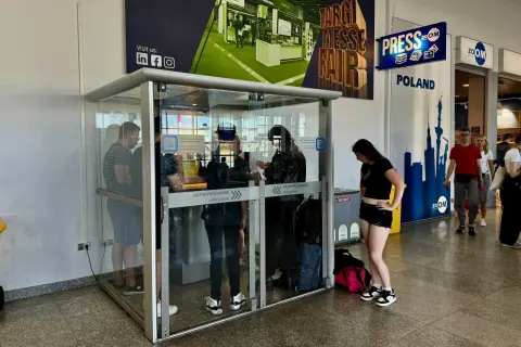 Szklana kabina na lotnisku w Poznaniu, oznaczona jako palarnia, z ludźmi w środku i na zewnątrz.