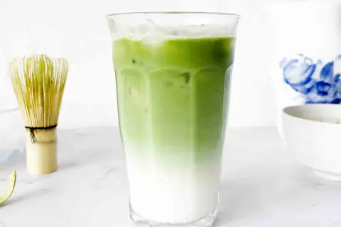 En snygg, isig matcha latte med lager av mjölk och grönt te. Bredvid står en visp för att göra matcha.