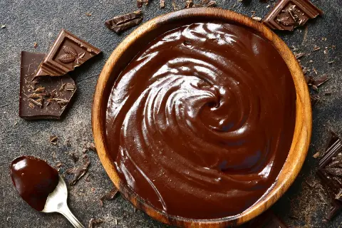 Un bol en bois déborde de chocolat fondu, prêt à travailler. Des éclats de chocolat et une cuillère en argent complètent cette scène gourmande.