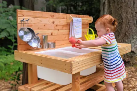 A toddler engages in Montessori movement activities, exploring a wooden mud kitchen with pots and a watering can.