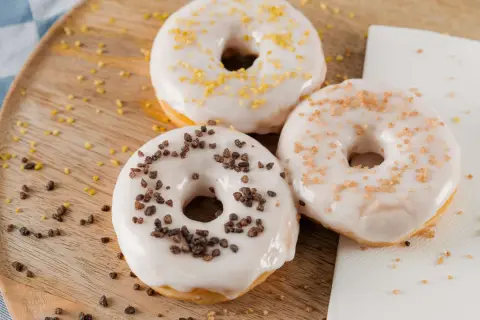 Drei Donuts mit weißer Glasur und Streuseln aus Schokolade, Zucker und Zitrone liegen auf einem Holzbrett.