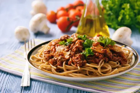 Une assiette de tagliatelles à la bolognaise, garnie de persil frais. Des champignons, tomates et une bouteille d'huile d'olive sont en arrière-plan. Une recette parfaite pour un repas réconfortant.