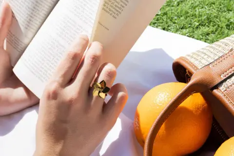Mano con un anillo dorado con forma de flor, leyendo un libro junto a naranjas y un bolso de mimbre. Las tallas de anillos perfectas para cada ocasión.