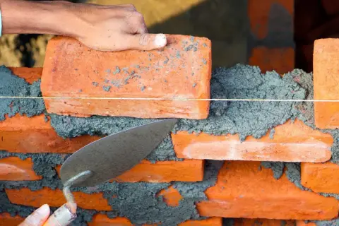Budowa muru z cegieł, gdzie cegły wystające co druga w murze tworzą wzór. Tynkarz układa kolejną cegłę.