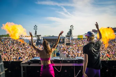 DJ-ka i DJ na scenie, tłum szaleje na festiwalu techno. Wybuchy ognia dodają energii.