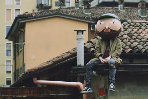 Un personaggio con una maschera gigante, simile a un cratere, siede sui tetti. I testi di Frah Quintale potrebbero raccontare la sua storia.