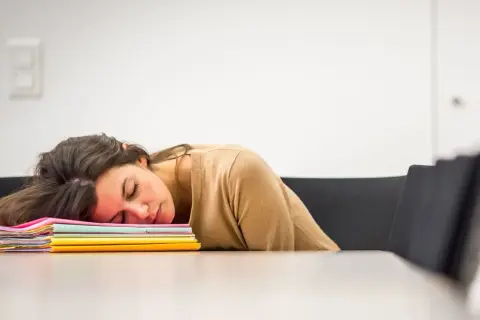 En kvinna sover uttråkad på jobbet, med huvudet vilande på en hög med färgglada mappar.