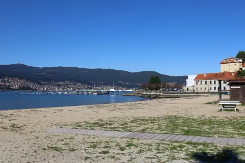 La playa de arealonga, con arena dorada y vegetación dispersa, se extiende hacia un mar azul tranquilo. Al fondo, un puerto deportivo con barcos y una ciudad en la ladera de una montaña verde.