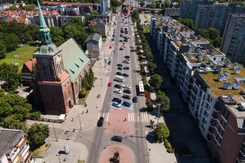 Widok z lotu ptaka na miasto z kościołem i długimi ogrodami. Ulicą jedzie autobus, a obok parkują samochody.