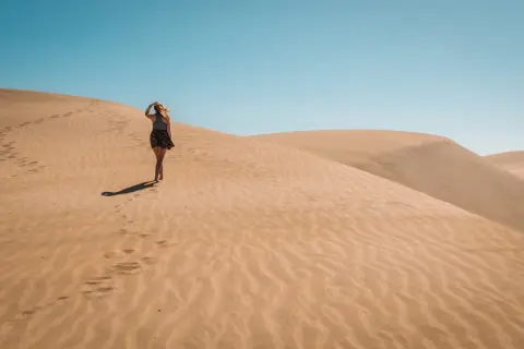 Kobieta spaceruje po piaszczystych wydmach Dunas de Maspalomas, zostawiając ślady na piasku pod błękitnym niebem.