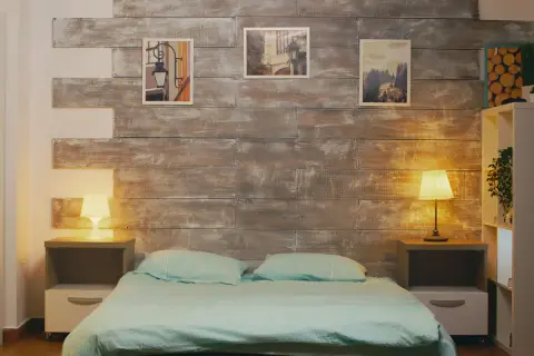 Przytulna sypialnia bez okna, z łóżkiem w turkusowej pościeli i drewnianą ścianą. Lampki nocne tworzą ciepły nastrój.