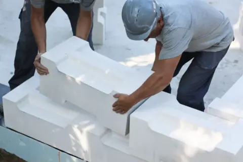 Dwóch budowlańców układa białe bloczki z perlitu, tworząc fundament. Jeden z nich w hełmie precyzyjnie dopasowuje element.