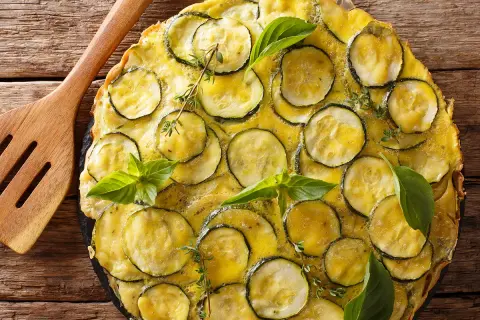 Une délicieuse scarpaccia de courgettes, garnie de basilic frais et de thym, prête à être servie avec une spatule en bois.