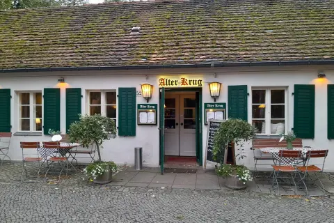Der "Alter Krug" in Dahlem, ein charmantes Gasthaus mit grünen Fensterläden und einem moosbewachsenen Dach, lädt mit seinen Tischen im Freien zum Verweilen ein.