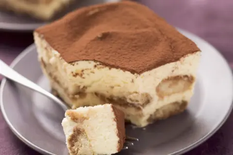 Ein Stück cremiges Tiramisu ohne Zucker, bestäubt mit Kakao, auf einem Teller mit einer Gabel.