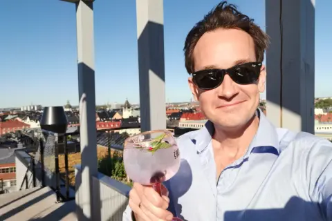 En man med solglasögon håller en rosa drink på en takterrass med utsikt över staden. En perfekt dag för en drink från arc blique by nobis.