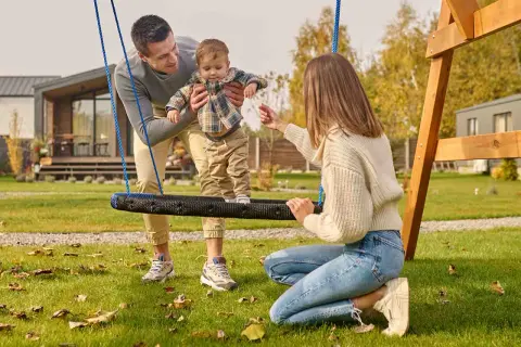 Rodzina cieszy się wspólnymi zabawami na podwórku. Tata trzyma dziecko na huśtawce, a mama zachęca do zabawy.