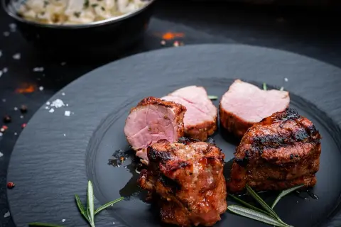 Saftige schweine medaillons, perfekt gegrillt, liegen auf einem dunklen Teller, garniert mit Rosmarin. Daneben eine Schüssel mit cremiger Beilage.