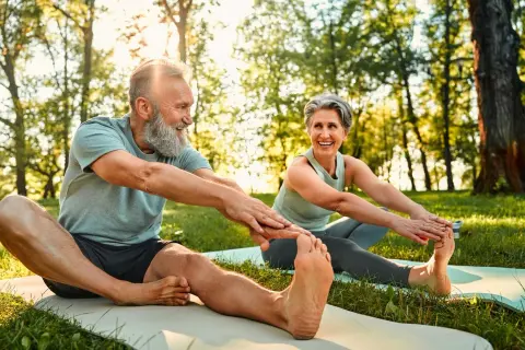Couple souriant pratiquant le qi gong en plein air, étirant leurs jambes sur des tapis de yoga dans un parc ensoleillé.