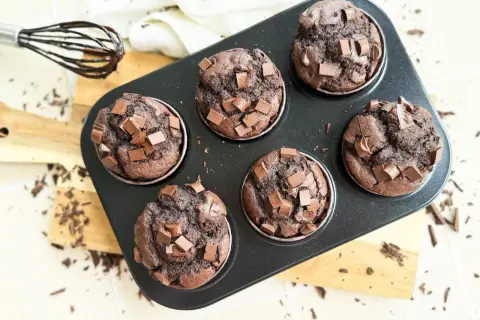 Sechs saftige, vegane Schokomuffins mit Schokostückchen in einer Muffinform. Ein Schneebesen mit Schokospuren liegt daneben.