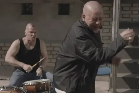 Dwóch mężczyzn, jeden gra na perkusji, drugi tańczy. To scena z teledysku zespołu Kult, który mieszka w Polsce.