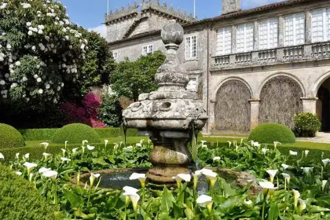 Jardín del Pazo de Oca con fuente de piedra y calas blancas, rodeado de setos y un gran rododendro en flor.