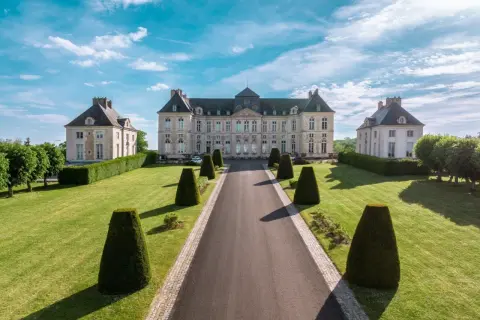 Le majestueux château de Brienne, avec ses ailes symétriques et son allée bordée de topiaires, sous un ciel bleu éclatant.