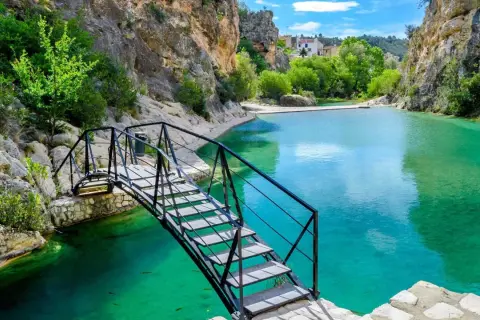 Puente metálico sobre aguas turquesas en uno de los lagos en Valencia, rodeado de rocas y vegetación.