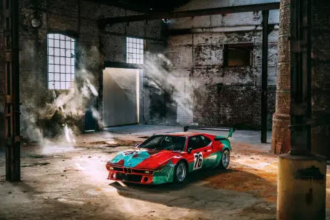 Un m1 coche de carreras con librea roja y verde, número 76, descansa en un almacén polvoriento con rayos de sol y humo.