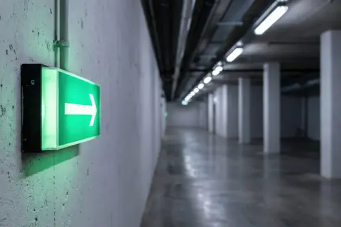 Zielone oświetlenie ewakuacyjne ze strzałką w prawo wskazuje drogę w długim, pustym korytarzu z betonowymi słupami.