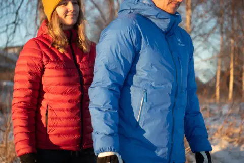 Två personer i vinterkläder, en i röd och en i blå, varmaste dunjackan, står utomhus i snö.