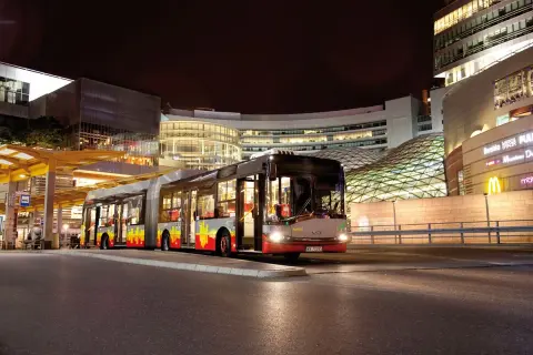 Długi, przegubowy autobus linii 59 w Łodzi nocą, zatrzymuje się przy nowoczesnym centrum handlowym.