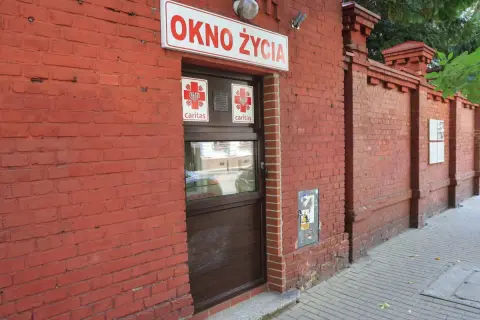 Czerwona ceglana ściana z napisem "OKNO ŻYCIA" i drzwiami. To miejsce daje szansę na nowe życie.