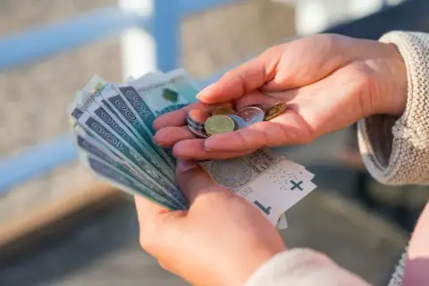 Dłonie trzymające banknoty 100 zł i monety. Może to być świadczenie kompensacyjne lub wypłata.