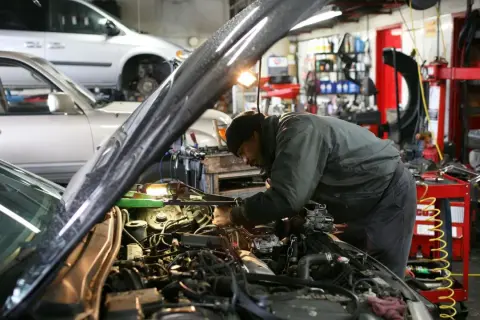 Mekaniker arbetar med en bilmotor i ett garage. En annan bil står i bakgrunden. Här erbjuds stor service.