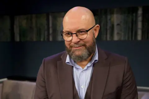 Szymon Bobrowski w garniturze i okularach, z brodą, wygląda jakby opowiadał o swoich filmach, serialach i programach.