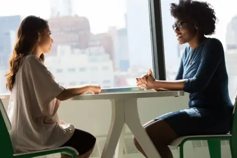 Dos mujeres conversan animadamente en una mesa junto a una ventana. Una de ellas, con gafas, sonríe mientras habla, demostrando su habilidad **speaking b1**.