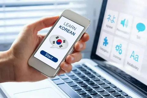 Dłoń trzyma smartfon z napisem "LEARN KOREAN" i flagą Korei. Obok laptop z ikonami do nauki języka koreańskiego od podstaw.
