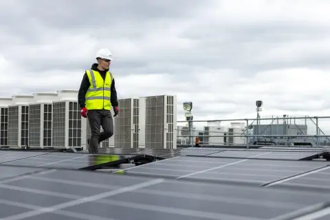 Pracownik w kamizelce odblaskowej przechodzi obok rzędu klimatyzatorów na dachu, gdzie instalowane są panele słoneczne. sofar solar polska.