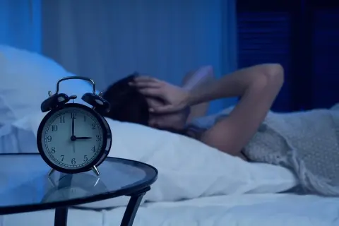 Budzik pokazuje 3:00. Osoba w łóżku, z rękami na głowie, cierpi z powodu problemów ze snem.