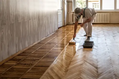 Un homme, masqué, utilise un rouleau pour vitrifier ou cirer un parquet en bois, lui redonnant son éclat.