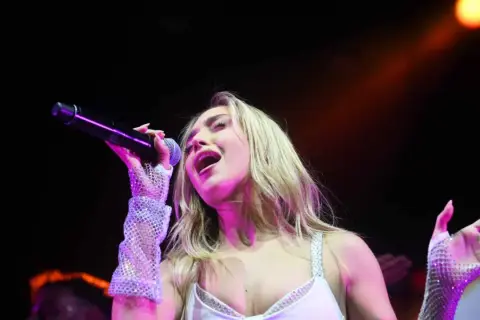 Ana Mena canta en Zaragoza, con un micrófono en la mano y guantes brillantes.