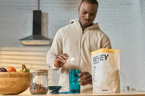 Mężczyzna przygotowuje odżywkę białkową po treningu. Na blacie stoi opakowanie WHEY PROTEIN, shaker i miska z owocami.