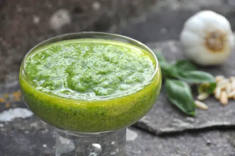 Pesto di basilico bimby in una coppetta, con aglio, pinoli e foglie di basilico sullo sfondo.