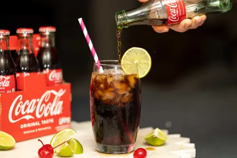 Coca-Cola nalewana do szklanki z lodem i limonką, obok wiśni i cytryn.