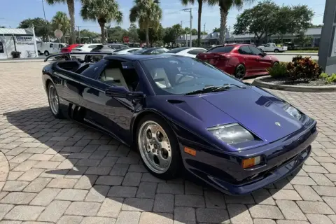 Fioletowy Lamborghini Diablo z jasnym wnętrzem, zaparkowany na brukowanym podjeździe, z palmami w tle.