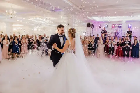 Para młoda tańczy na weselu, goście patrzą, zespół gra. W tle konfetti i dym. Piękny moment, ale czy wiesz o ukrytych kosztach wesela?