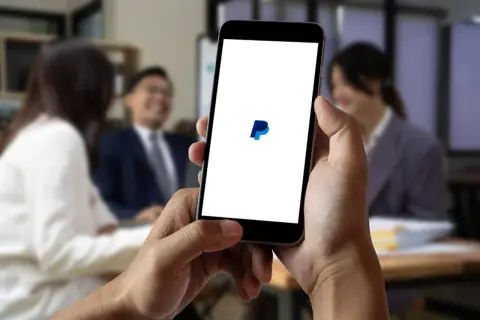 Dłoń trzyma smartfon z logo PayPal na białym ekranie, w tle rozmyte osoby w biurze.