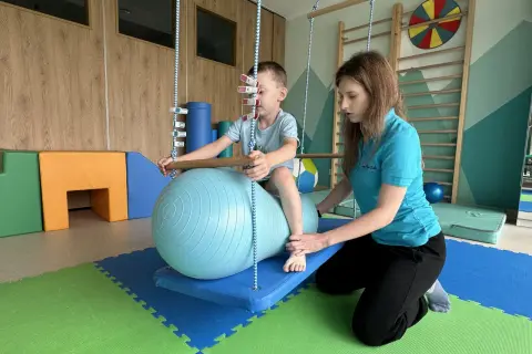 Terapeuta pomaga dziecku na huśtawce z piłką terapeutyczną w sali integracji sensorycznej.